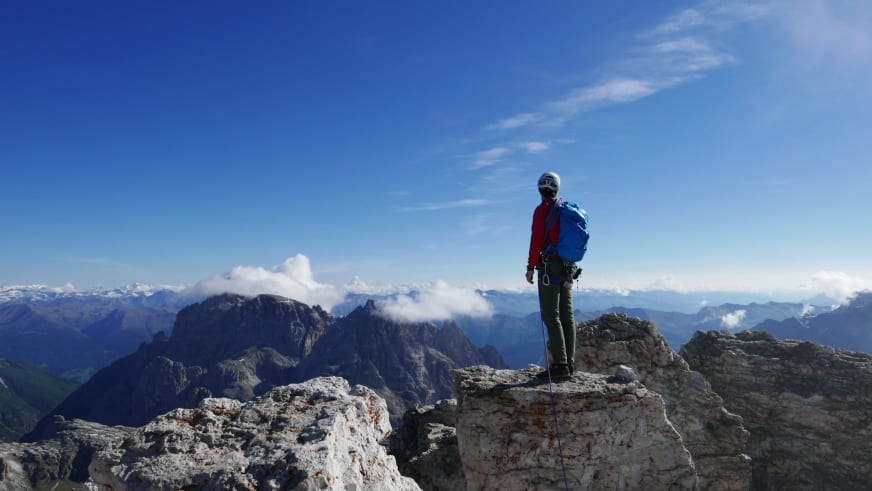 Auf hellem Kalksteingipfel steht Bergführerin Matthias Larcher mit Klettergurt und rotem Seil und überblickt das Bergpanorama.