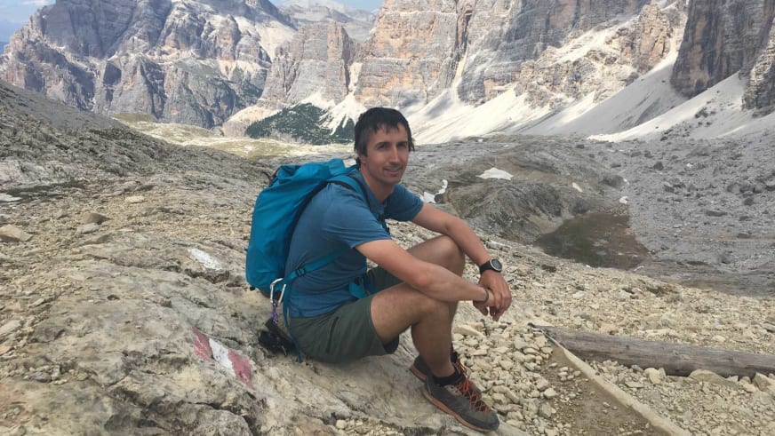 In blauem T-Shirt und grünen Shorts sitzt Bergführerin Matthias Larcher entspannt auf Felsvorsprung vor alpiner Karstlandschaft.
