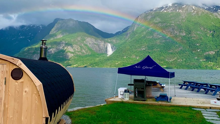 Holz-Fasssauna vom Nes Gard direkt am Fjordufer mit Regenbogen über der Bergkulisse im Hintergrund.