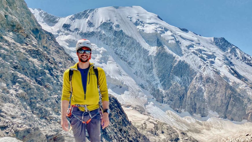 Bergführer Jakob Halder in gelber Softshelljacke und weißem Helm lächelt vor schneebedecktem Berggipfel.