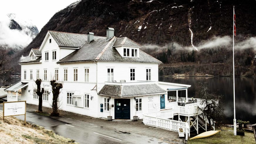 Weißes Holzgebäude des Fjærland Fjordstove Hotells direkt am Ufer des Sognefjords mit schneebedeckten Bergen im Hintergr