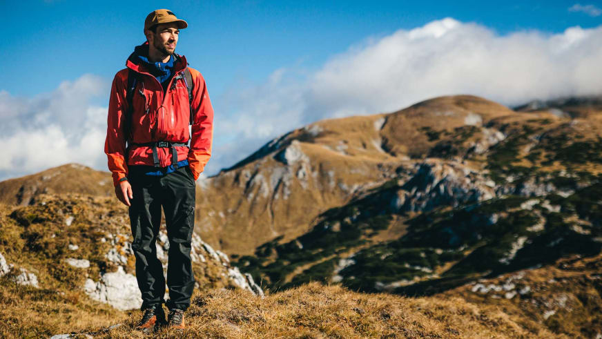 In roter Outdoorjacke und brauner Kappe steht Bergführer Jakob Halder auf gelblichem Grasland vor Berghängen.