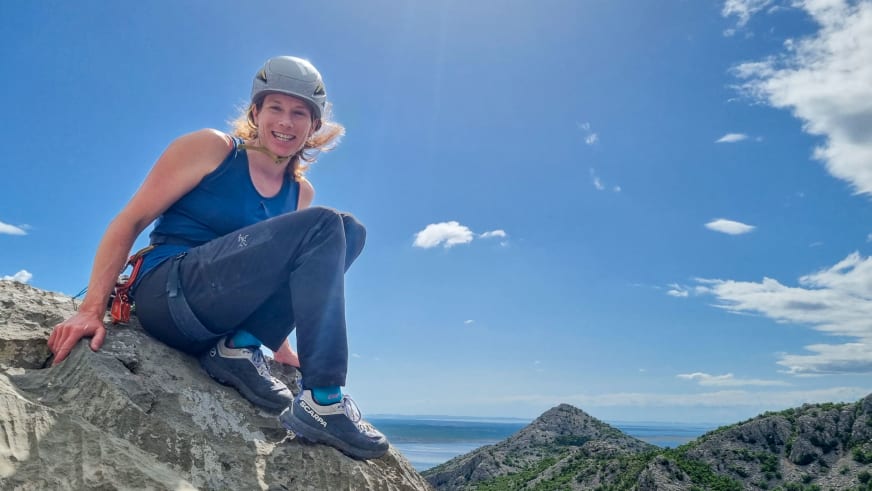 Auf hellem Felsvorsprung sitzt Bergführerin Tanja Kuster mit weißem Helm und Klettergurt vor Küstenlandschaft.