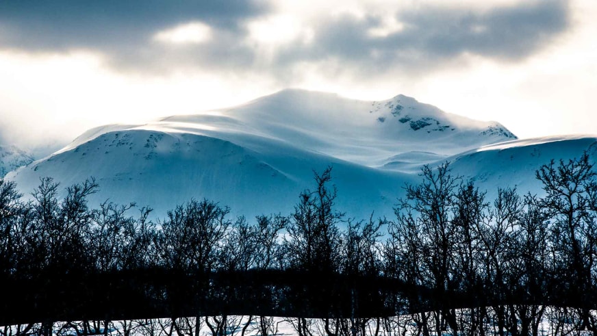 Bergführer Felix Gruber zeigt den schneebedeckten Fastdalstinden in Lyngen mit gerundeten Gipfeln unter bewölktem Himmel.