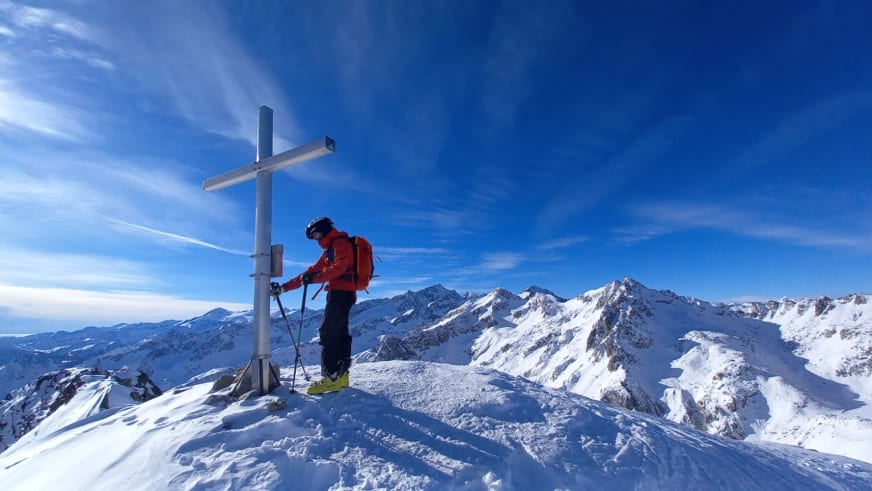 Am verschneiten Gipfel steht Bergführer Cesare Lotti neben weißem Gipfelkreuz mit Skistöcken.