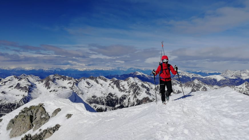 Auf verschneitem Gipfel steht Bergführer Cesare Lotti mit Tourenskiern vor Alpenpanorama.