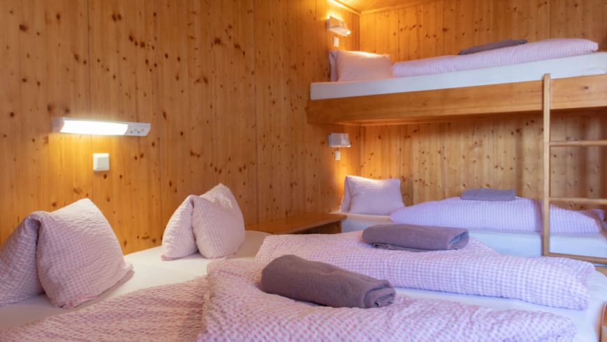 Mehrbettzimmer in der Schihütte Zams mit Doppelbett und Etagenbett aus hellem Holz und karierter Bettwäsche.