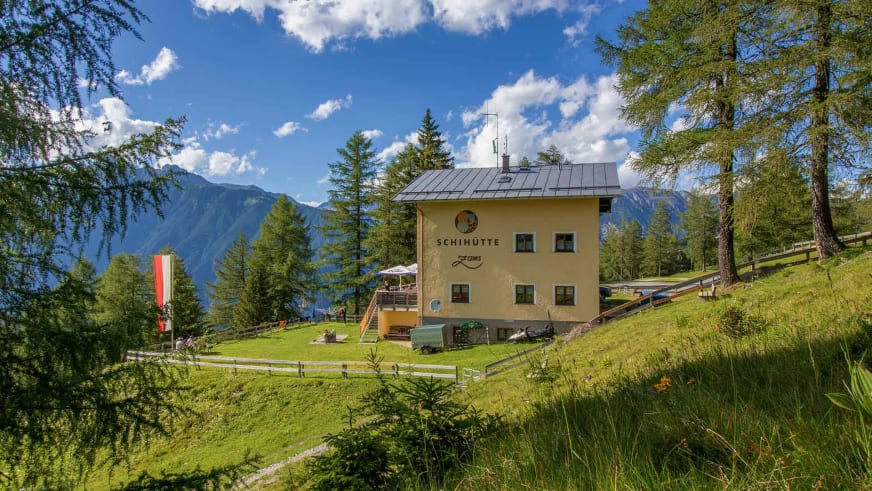 Außenansicht der Schihütte Zams mit gelber Fassade und Terrasse auf grüner Almwiese in den Bergen.