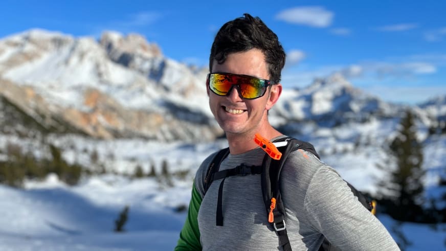 Bergwanderführer Patrick Hindelang mit goldener Sonnenbrille und schwarzem Rucksack vor verschneiter Berglandschaft in den Dolomiten.