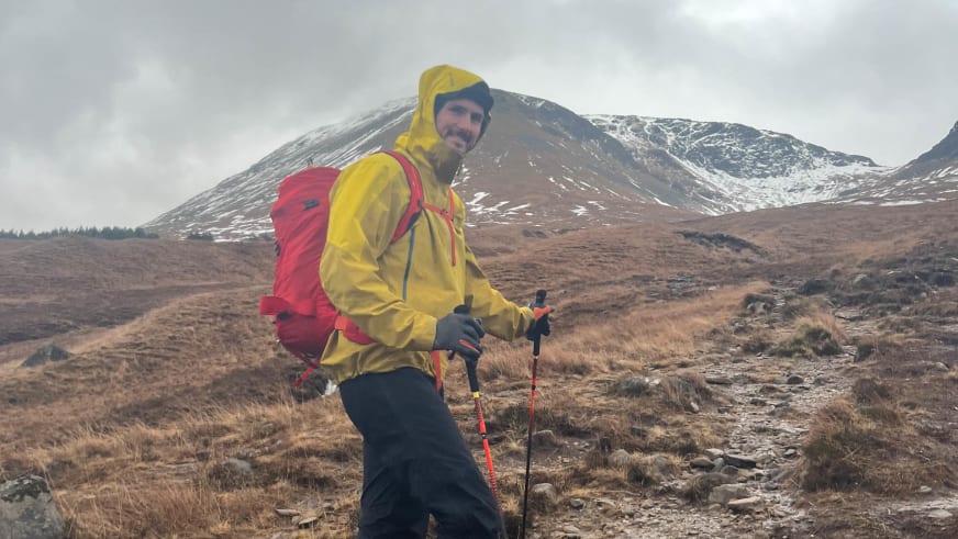 Bergwanderführer Patrick Hindelang in gelber Regenjacke mit roten Wanderstöcken im schottischen Moorland vor schneebedeckten Bergen.