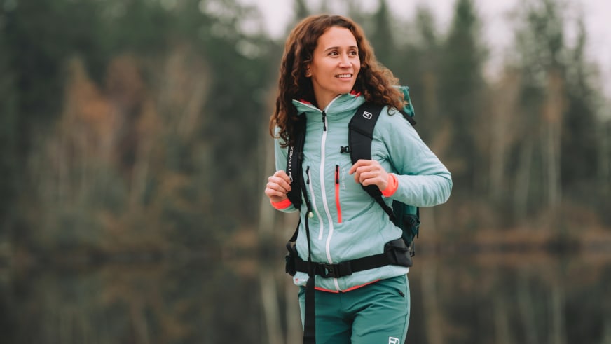 Bergwanderführerin Corina Machon trägt hellblaue Softshelljacke und schwarzen Rucksack vor grünem Waldhintergrund.