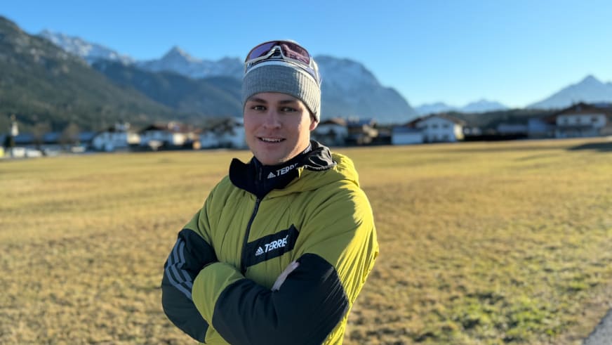 Bergführer Robert Kadner trägt gelb-schwarze Daunenjacke und graue Mütze vor verschneiter Alpenlandschaft mit Ortschaft.