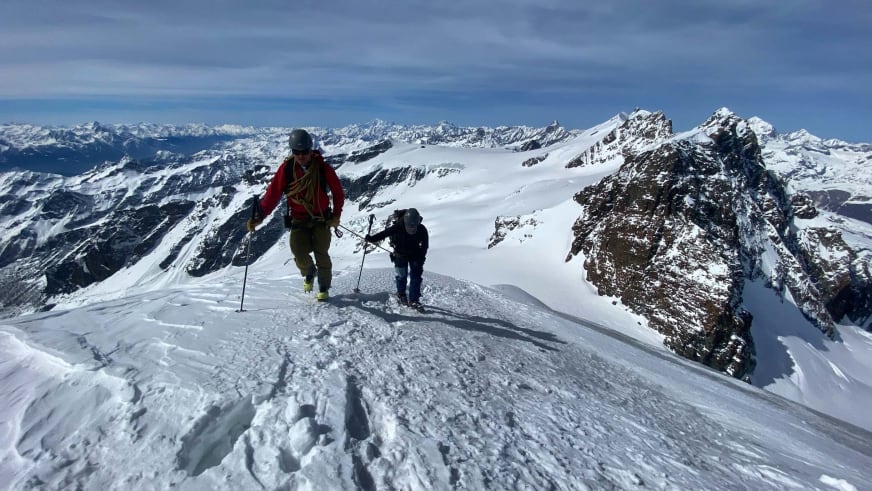 Zwei Personen gehen auf verschneitem Grat aufwärts, Bergführer Robert Kadner führt die Hochtour mit Helm und Bergstöcken.