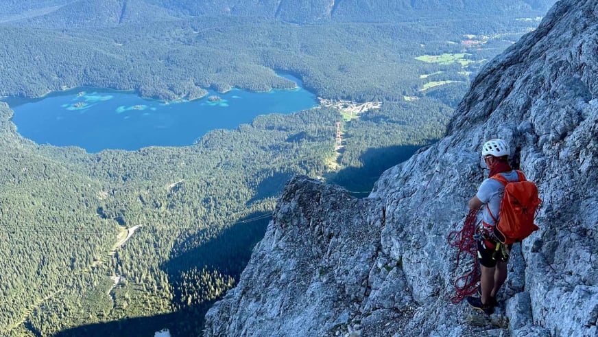 An grauer Felswand steht Bergführer Robert Kadner mit rotem Kletterseil und weißem Helm über Seenlandschaft im Tal.