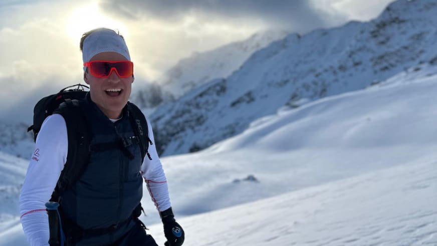 Lächelndes Portrait von Bergführer Robert Kadner in grauer Mütze und roter Skibrille vor verschneiter Berglandschaft.