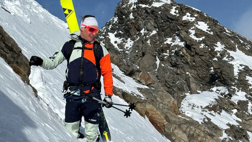 Bergführer Robert Kadner steht mit Skistöcken und gelbem Ski am steilen Schneehang vor massiver Felswand bei Sonnenschein.