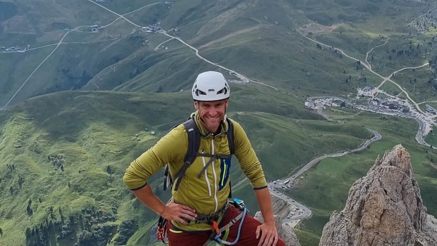 Bergführer Josef Mayr mit weißem Helm und gelbem Softshell-Oberteil lächelt am Felsgipfel mit Blick ins grüne Tal.