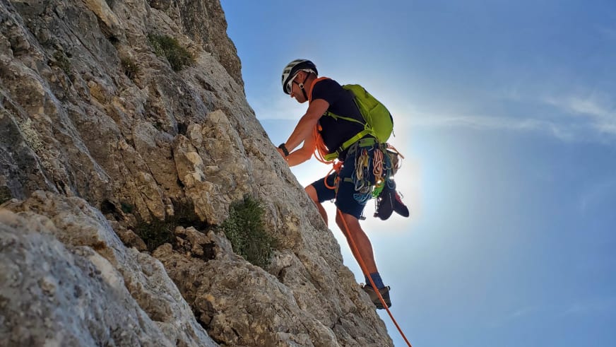 An steiler Kalksteinwand klettert Bergführer Josef Mayr mit weißem Helm und grünem Rucksack bei Sonnenschein.