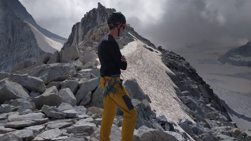 Bergführerin Luca Trubbiani in gelber Berghose und grünem Helm geht über ein Geröllfeld aus grauen Steinblöcken.