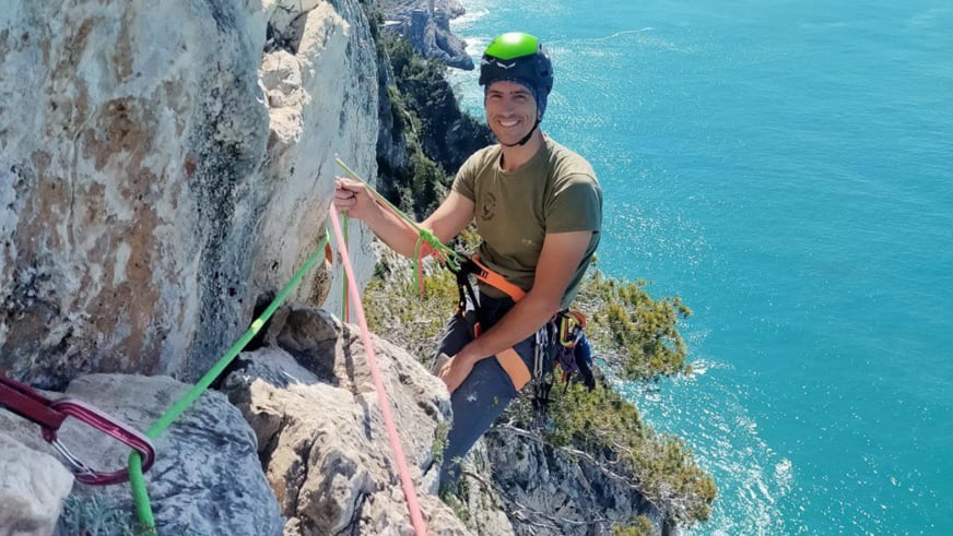 An einer steilen Felswand über türkisblauem Meer sitzt Bergführerin Luca Trubbiani mit Kletterseilen.