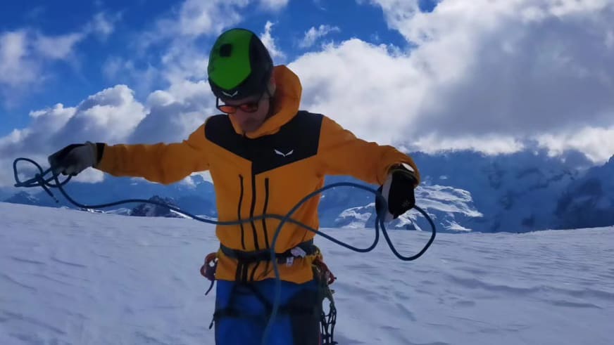 Bergführerin Luca Trubbiani in oranger Bergjacke hält ein schwarzes Seil auf einer weiten Schneefläche in den Händen.