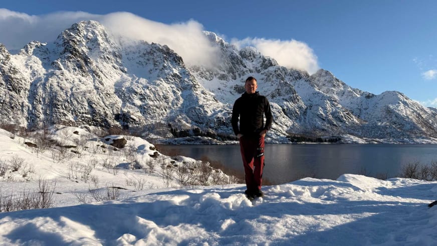 Portrait zeigt Bergführer Wolfgang Pusch in schwarzer Winterjacke und roten Hosen vor einer verschneiten Fjordlandschaft.