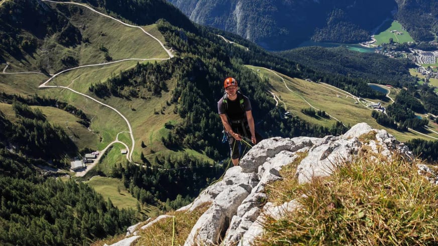 Auf einem Kalksteingrat hält Bergführer Wolfgang Pusch mit orangem Helm ein grünes Seil über einem Alpental.