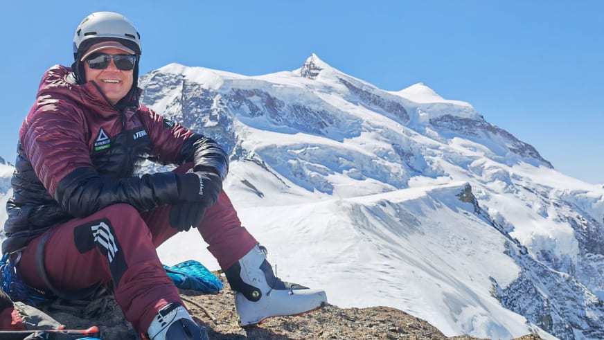 Bergführer Wolfgang Pusch sitzt in bordeauxroter Daunenjacke und weißem Helm vor einem schneebedeckten Viertausender.