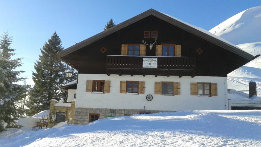 Die Schönfeldhütte zeigt sich im Winter mit verschneiter Alpenarchitektur und Hirschgeweih am Giebel.