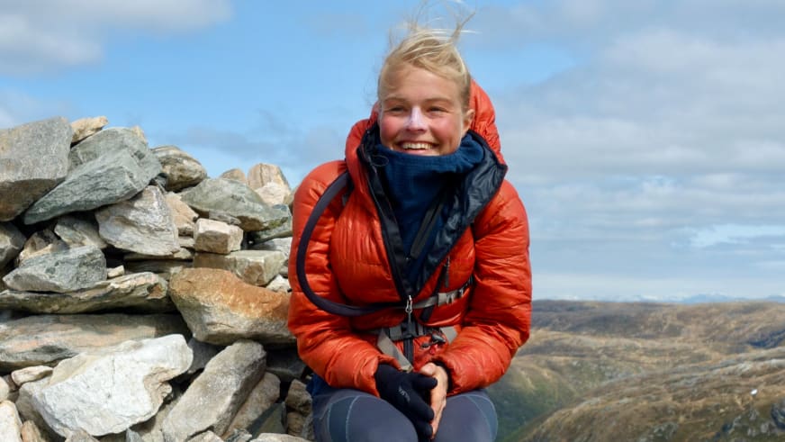 In orangefarbener Daunenjacke sitzt Bergwanderführerin Nora Coenen lächelnd neben einem aufgeschichteten Steinturm.