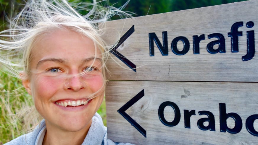Bergwanderführerin Nora Coenen lächelt vor einem hölzernen Wegweiser mit blonden, vom Wind bewegten Haaren.