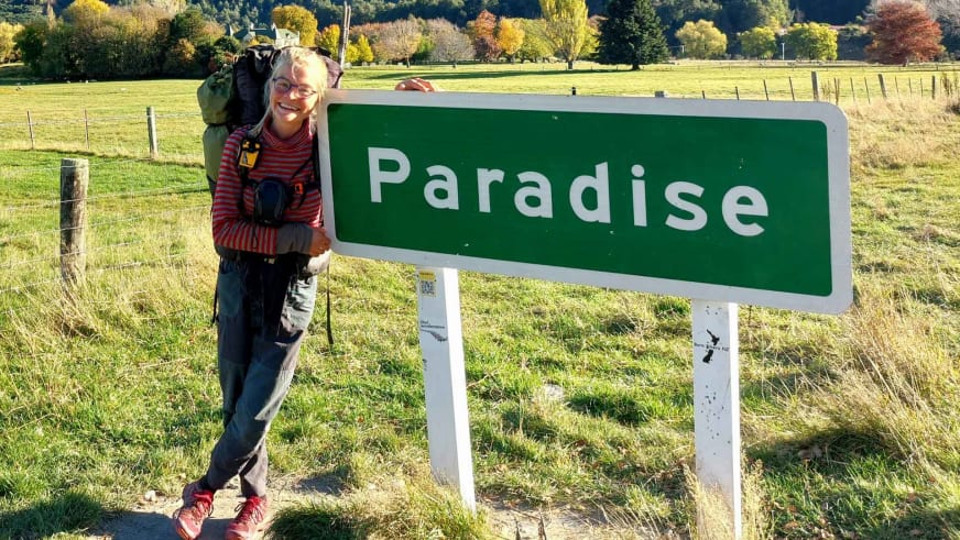 Neben einem grünen Paradise-Ortsschild steht Bergwanderführerin Nora Coenen mit Wanderausrüstung und grünem Rucksack.