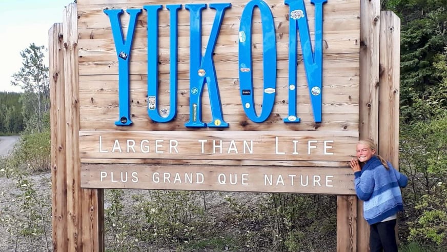 Bergwanderführerin Nora Coenen in blauer Fleecejacke hält sich am hölzernen Yukon-Ortsschild fest und lächelt in die Kamera.