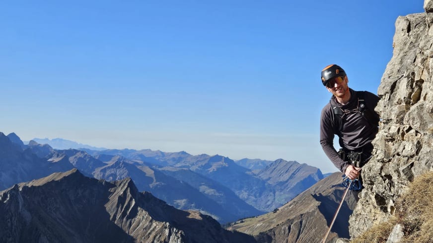 An hoher Bergwand steht Bergführer Lukas Degen mit Klettergurt und Seil vor weitem Alpenpanorama.