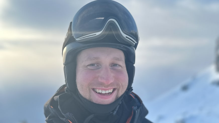 Lächelnder Bergführer Lukas Degen mit schwarzem Helm und Skibrille vor verschneiter Bergkulisse im Winter.