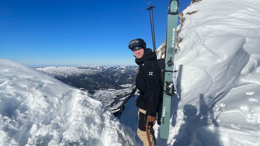 Bergführer Lukas Degen steht lächelnd mit grünen Tourenski auf verschneitem Gipfel vor Winterlandschaft.