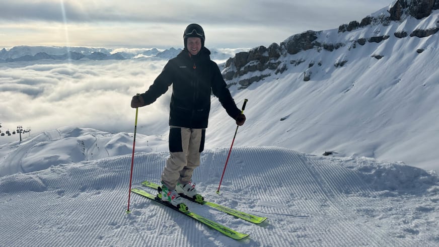 Bergführer Lukas Degen steht lächelnd auf grünen Skiern im Pistenschnee über einer geschlossenen Wolkendecke.