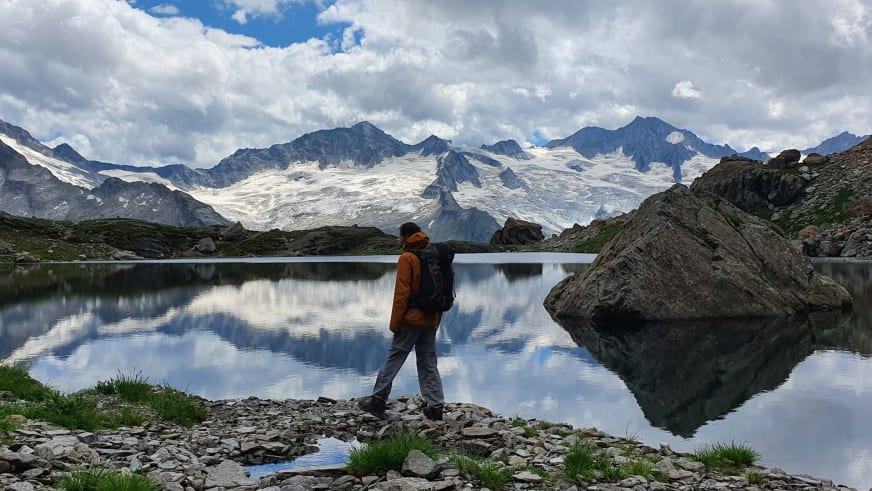 Bergwanderführer Jonas Bergmann in oranger Jacke steht am steinigen Ufer eines Bergsees vor schneebedeckter Gebirgskette.