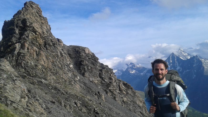 Bergwanderführer Jonas Bergmann mit grauem Rucksack und hellblauem Shirt vor hoher Felswand aus grauem Gestein.
