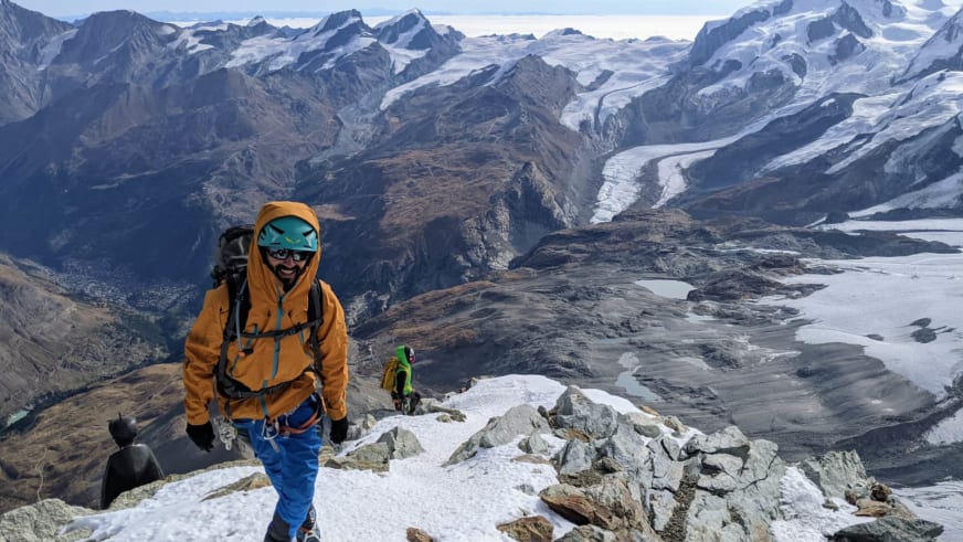 Bergwanderführer Jonas Bergmann in oranger Jacke steht mit zweiter Person auf schneebedecktem Felsgrat vor Gletscherlandschaft.