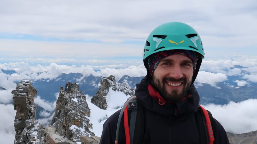 Lächelnder Bergwanderführer Jonas Bergmann mit türkisem Kletterhelm und schwarzer Jacke auf hochalpinem Gipfel über Wolkendecke.