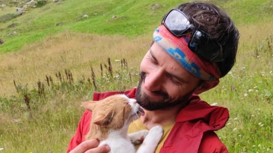 Bergwanderführer Jonas Bergmann mit roter Jacke und buntem Stirnband hält weiß-braune Katze auf grüner Bergwiese.