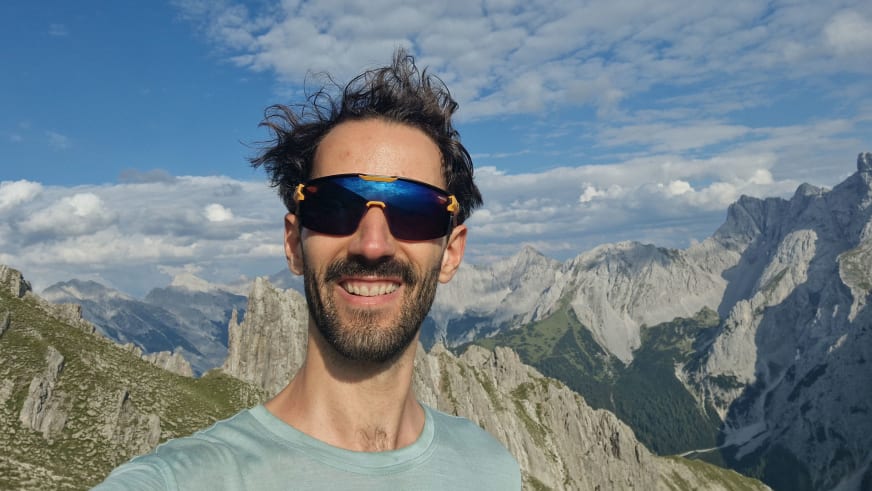 Bergwanderführer Jonas Bergmann mit Sonnenbrille und hellblauem T-Shirt vor alpiner Kalksteinlandschaft mit schroffen Felsgipfeln.