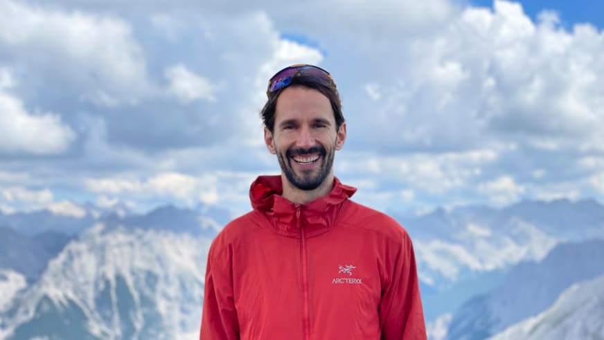 Bergwanderführer Jonas Bergmann mit Vollbart lächelt in roter Arc'teryx Jacke vor verschneiter Bergkulisse bei Tageslicht.