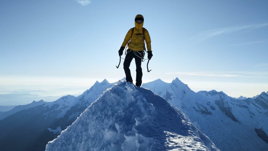 Bergführer Alexander Brückler steht auf verschneitem Berggrat mit zwei Eisgeräten in gelber Daunenjacke vor schneebedeckten Gipfeln.