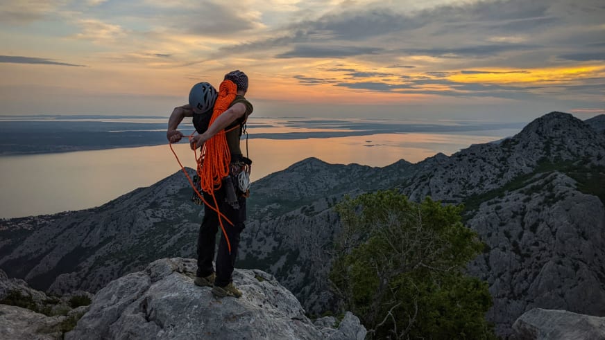 Bergführer Alexander Brückler organisiert orangefarbenes Kletterseil auf Felsvorsprung mit Küstenlandschaft im Hintergrund.