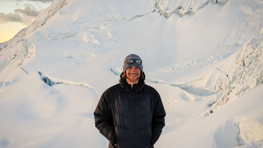 Bergführer Alexander Brückler lächelt vor verschneiter Gletscherlandschaft und trägt schwarze Daunenjacke mit grauer Mütze.