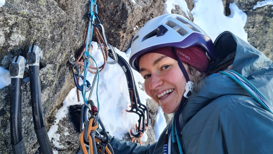 Bergwanderführerin Magdalena Strötz macht ein Selfie mit weißem Kletterhelm und blauer Daunenjacke vor grauem Fels mit Schneeresten.