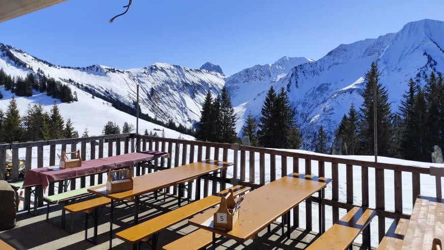 Holzterrasse des Neuhornbachhaus mit Biergarnituren und Panoramablick auf verschneite Gipfel der Allgäuer Alpen.