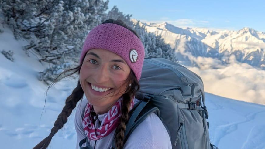 Bergführerin Monika Hümmer lächelt mit violetter Mütze vor verschneiter Bergkulisse.